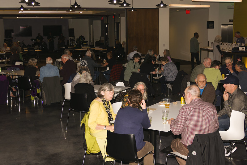Blues Jam-goers are seated around several tables, looks like some animated conversations, and there are musical instruments in the background.