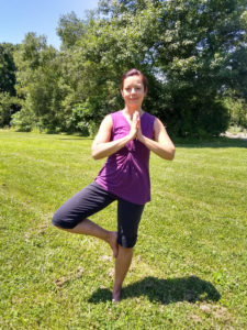 Woman wearing purple top and gray yoga pants in a pose in an outdoor setting