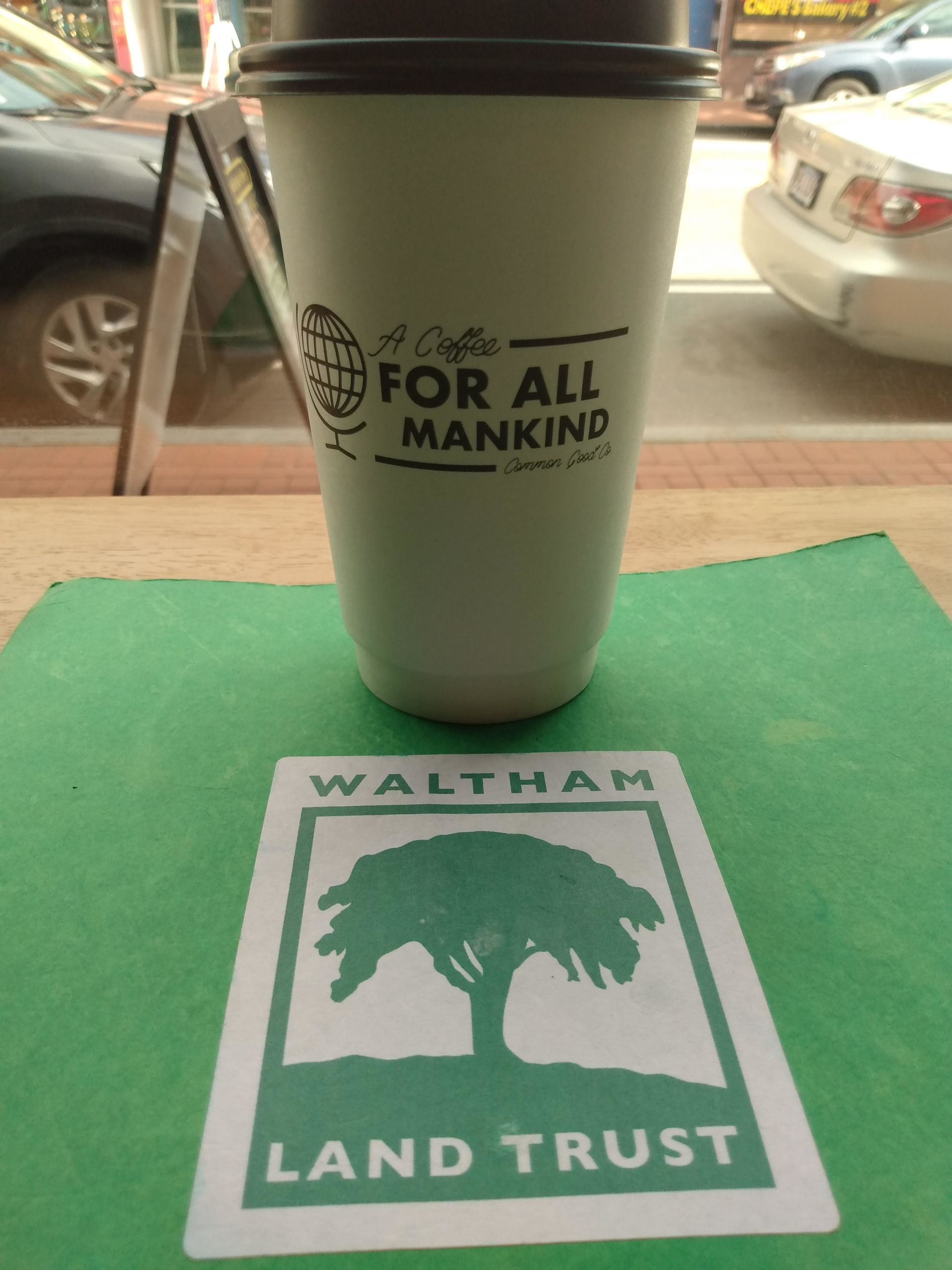 Common Good cafe coffee mug is placed on a table above an image of the Waltham Land Trust Logo